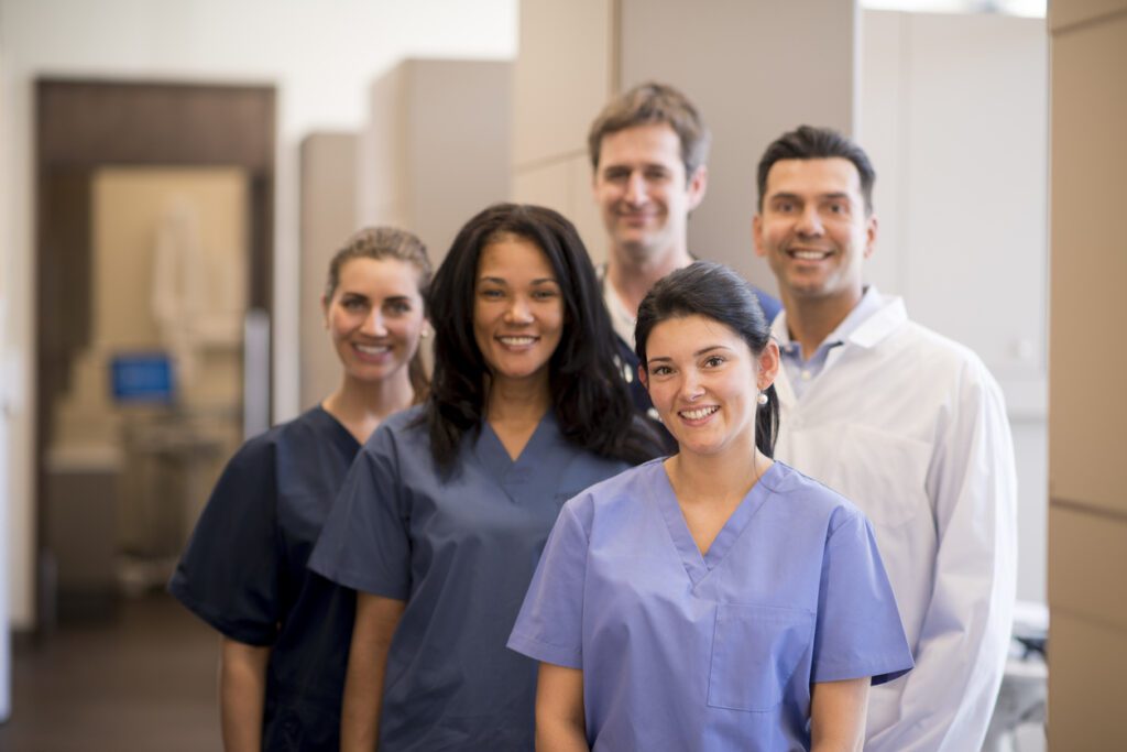 Adiverse team of smiling medical professionals in scrubs and a white coat standing in a modern clinic hallway.